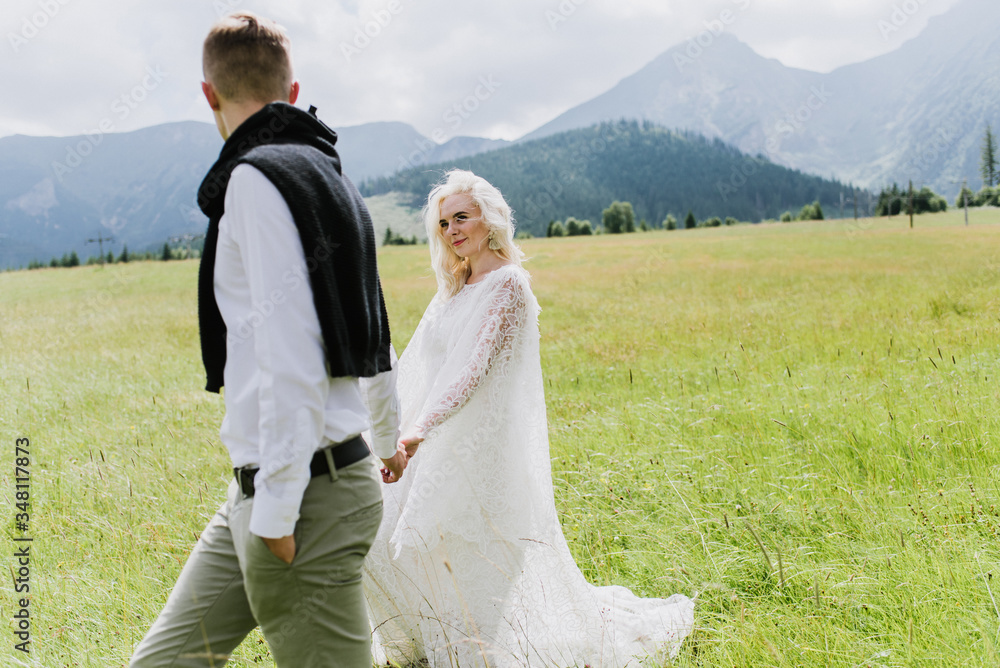 Beautiful bride in a boho style dress and groom walk on the field near the mountains. Wedding photo shoot in the mountains.