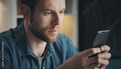 Close-up of young unshaven man in denim shirt holding cellphone with both hands and texting, home