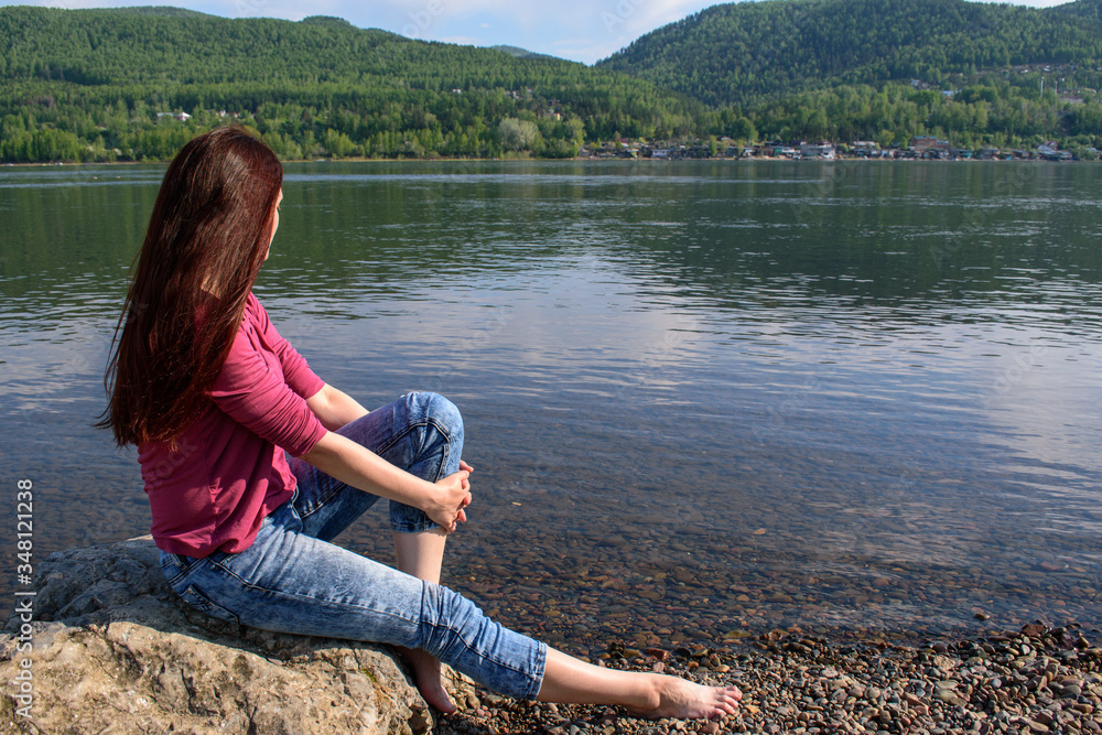 Woman on banks of a large river or lake. Long brown hair. Sits on a stone, profile view, face not visible.  Mountains, sky. Red jacket, jeans on ankle pad. Concept of summer vacation, thoughtfulness.