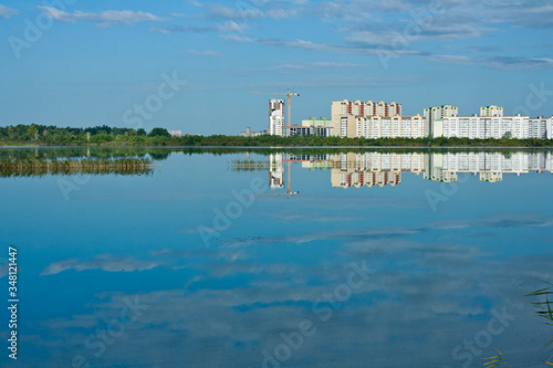 A clean lake with reeds, a blue cloudy sky and the construction of high-rise buildings in the background.