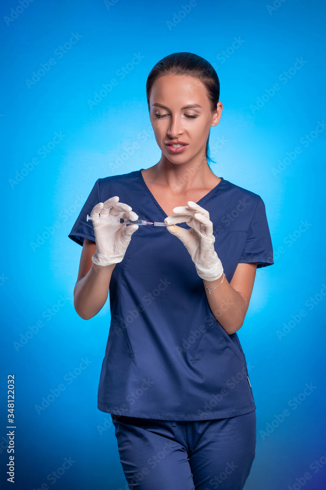 Brunette with a braided tail, posing while standing on a blue background, in a blue surgical suit, with a protective mask on his face, in medical gloves, wearing a cap from a syringe. Medical worker. Brunette with a braided tail, posing while standing on a blue background, in a blue surgical suit, with a protective mask on his face, in medical gloves, wearing a cap from a syringe. Medical worker.