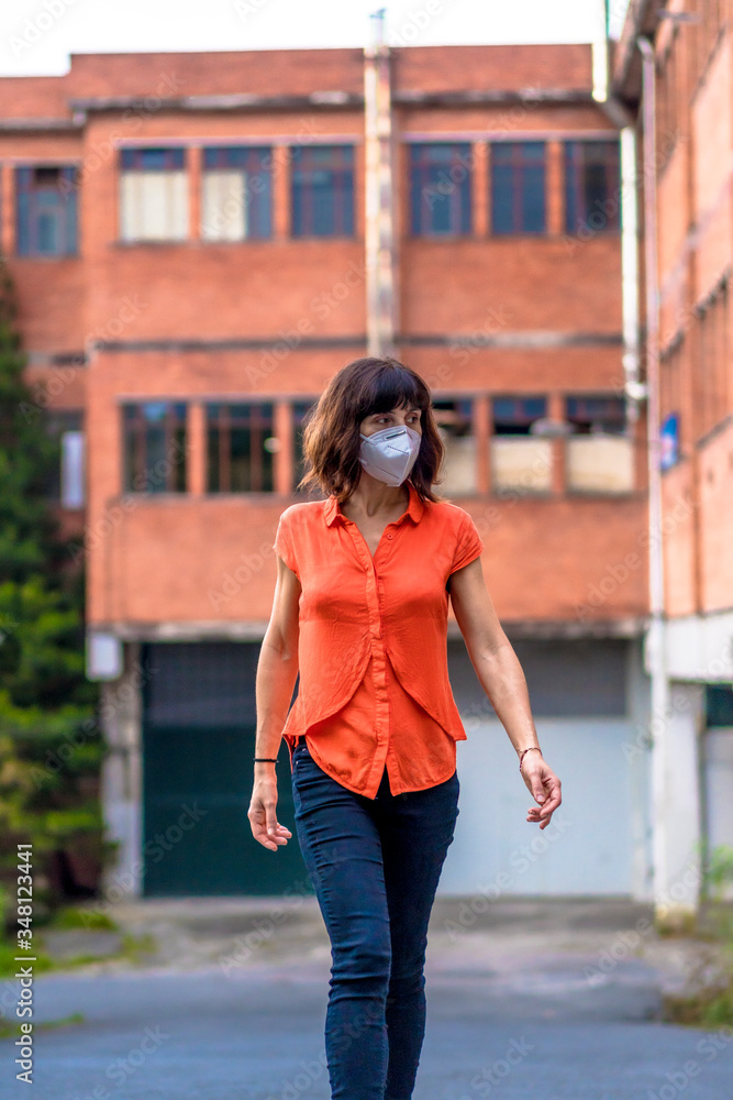 A young woman in a mask, orange shirt and black pants walking through the industry. Health emergency, pandemic Covid-19