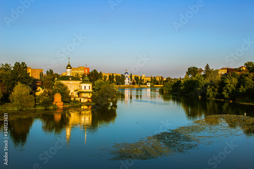 Summer city landscape. A river among urban buildings. Trees and buildings are reflected in the water.