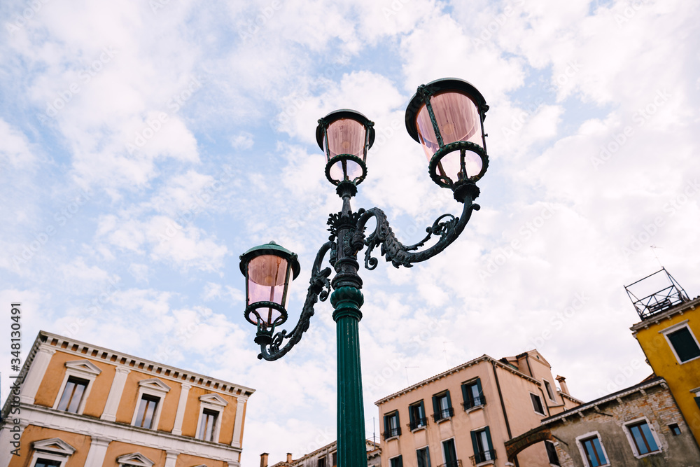 An ancient triple metal street lamp, with pink plafonds against the sky and buildings of Venice, Italy. Vintage street lighting, elegant pillar design.