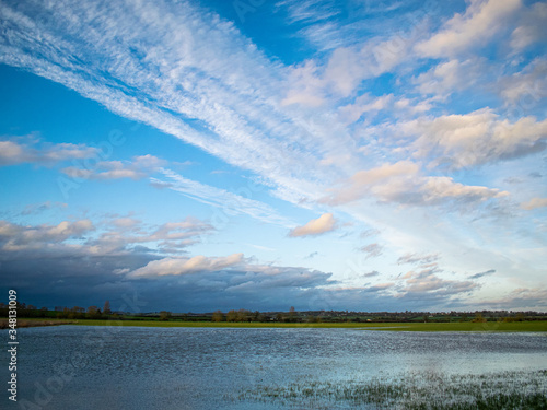 Rural landscape with dramatic cloudy sky, farm land floded by local river, blue sky with different types of clouds, birds sitting in the water, darker clouds approaching from far distance. photo
