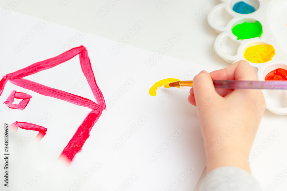 Child is drawing the sun and red house with watercolors on the white sheet of paper. Child is drawing the sun and red house with watercolors on the white sheet of paper.