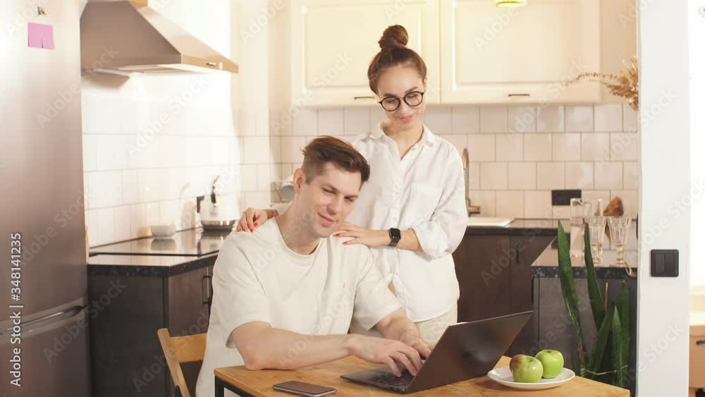 Beautiful caucasian married couple together at home, man work from home, at freelance. Careful wife support and hug him from back. Man use laptop, surf the Net Beautiful caucasian married couple together at home, man work from home, at freelance. Careful wife support and hug him from back. Man use laptop, surf the Net