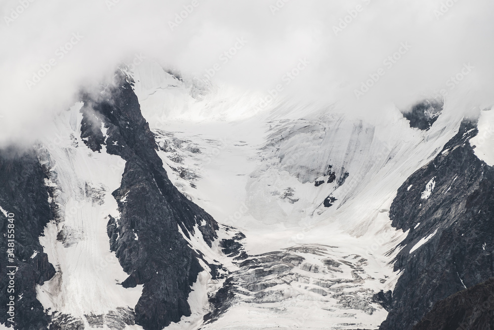 Obraz premium Atmospheric minimalist textured alpine landscape with massive glacier on big mountain in low clouds. Background of snowbound mountainside. Cracks on ice. Majestic foggy misty scenery on high altitude.
