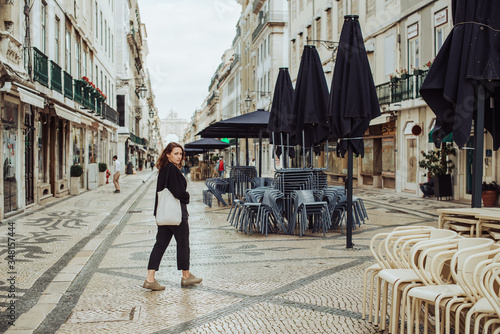 Millennial woman walking in the street by a closed restaurant during Covid-19 pandemic in Lisbon, Portugal. Looking back at camera. Empty touristic street