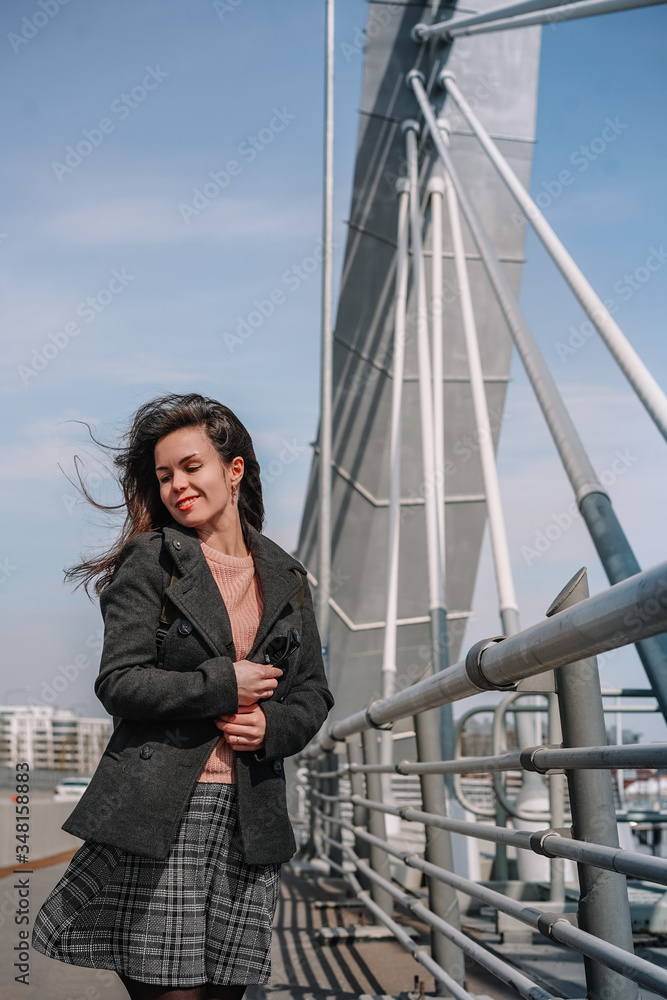 A young brunette girl in a coat and skirt stands on a bridge, beautiful metal structures in the air, hair flying in the wind, business issues, construction engineering A young brunette girl in a coat and skirt stands on a bridge, beautiful metal structures in the air, hair flying in the wind, business issues, construction engineering