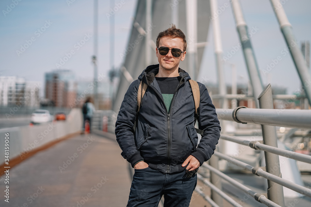 A young man in a jacket and glasses stands on a bridge, beautiful metal structures in the air, hair flying in the wind, business issues, construction engineering A young man in a jacket and glasses stands on a bridge, beautiful metal structures in the air, hair flying in the wind, business issues, construction engineering