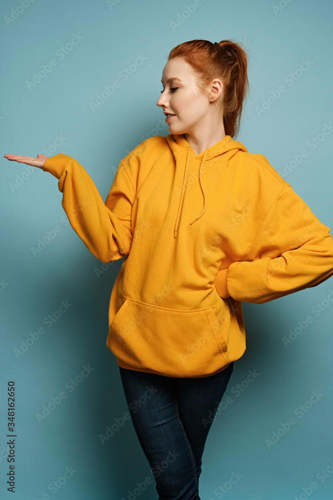 The red-haired girl stands on a blue background in a yellow hoodie, pointing her palm to the side and turning her head profile The red-haired girl stands on a blue background in a yellow hoodie, pointing her palm to the side and turning her head profile