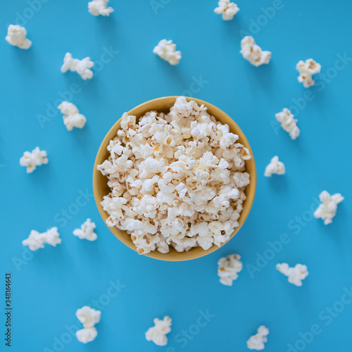 Popcorn in a yellow cup on a blue background and popcorn spread out nearby. Top view.