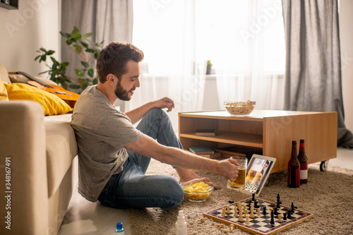 Positive bearded man sitting on floor and drinking beer with friend while communicating online in home isolation photo