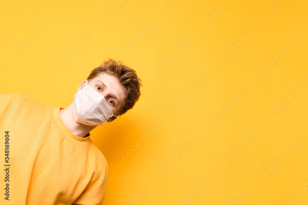 Funny guy in a medical mask on his face looks from the right side and looks at the camera, isolated on a yellow background. Coronavirus pandemic. COVID-19. Quarantine.