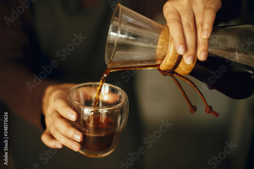 Professional female barista in black uniform making drip coffee. Person pouring fresh aromatic coffee from glass flask in cup, hands close up. Alternative coffee brewing, v60.