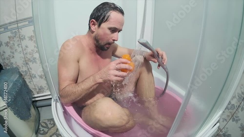 Adult man with beard sits in the pink tiny bath and plays with toy. Man goes bananas in the tiny bathroom.