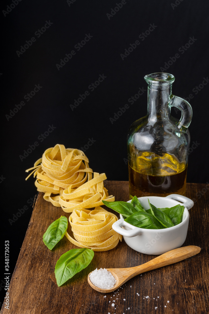 Aerial view of wooden spoon with salt, basil in white bowl, bottle of oil and raw tagliatelle, with selective focus, on wooden table, black background, vertical Aerial view of wooden spoon with salt, basil in white bowl, bottle of oil and raw tagliatelle, with selective focus, on wooden table, black background, vertical
