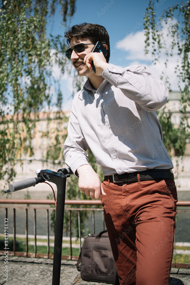 Portrait of guy in elegant clothes speaking on the phone. Happy boy with moustaches wearing sunglassesand talking on mobile while leaning on electric scooter. Portrait of guy in elegant clothes speaking on the phone. Happy boy with moustaches wearing sunglassesand talking on mobile while leaning on electric scooter.