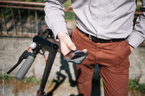 Cropped man holding smartphone while standing. Male hand holding black mobile phone while leaning on scooter rudder outdoor.