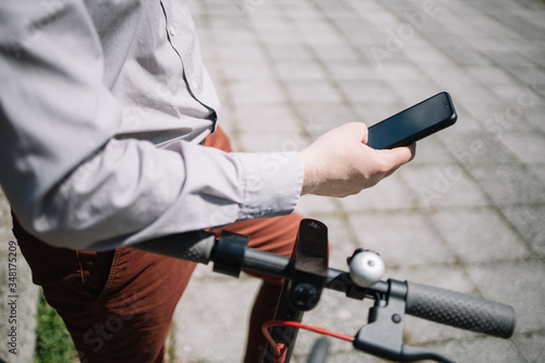 Close-up of man's hand holding black mobile phone. Side view of cropped man holding smartphone and leaning on scooter rudder in sunny day.