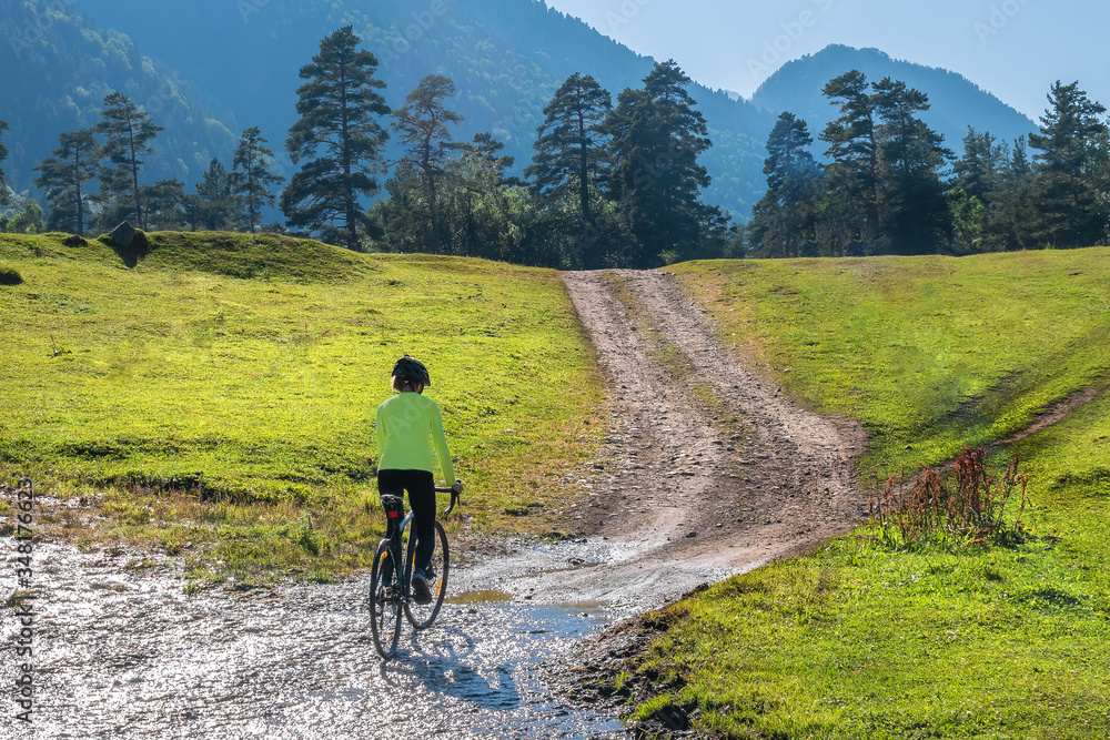 A young beautiful smiling girl on a cyclocross bike goes up overcoming difficulties on a green meadow against the background of forests and mountain peaks