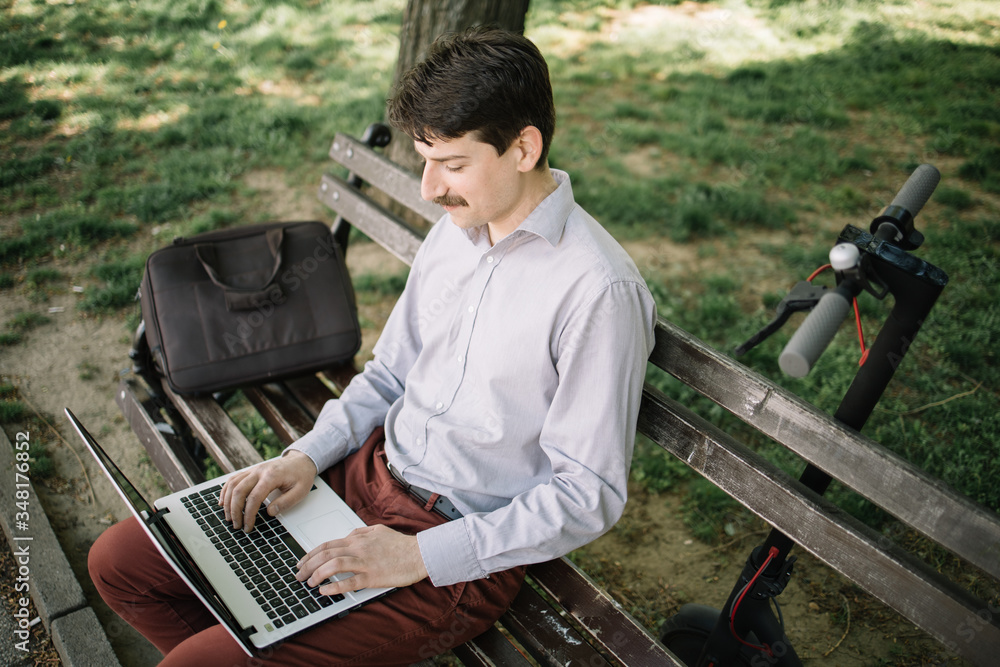 High angle view of man with laptop in nature. Guy with moustache wearing elegant outfit working on computer while sitting on bench near his bag. High angle view of man with laptop in nature. Guy with moustache wearing elegant outfit working on computer while sitting on bench near his bag.