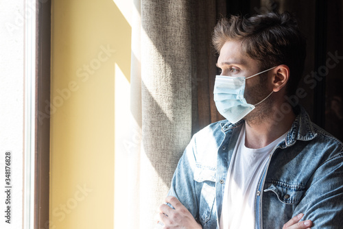 Man in medical mask with crossed arms looking at window in living room