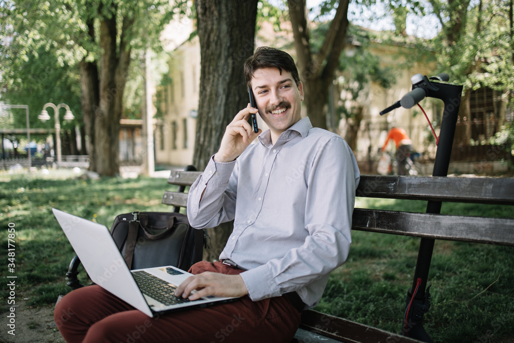 Happy man sitting on bench with laptop and holding mobile phone. Side view of smiling man with laptop on his lap speaking on the phone. Happy man sitting on bench with laptop and holding mobile phone. Side view of smiling man with laptop on his lap speaking on the phone.