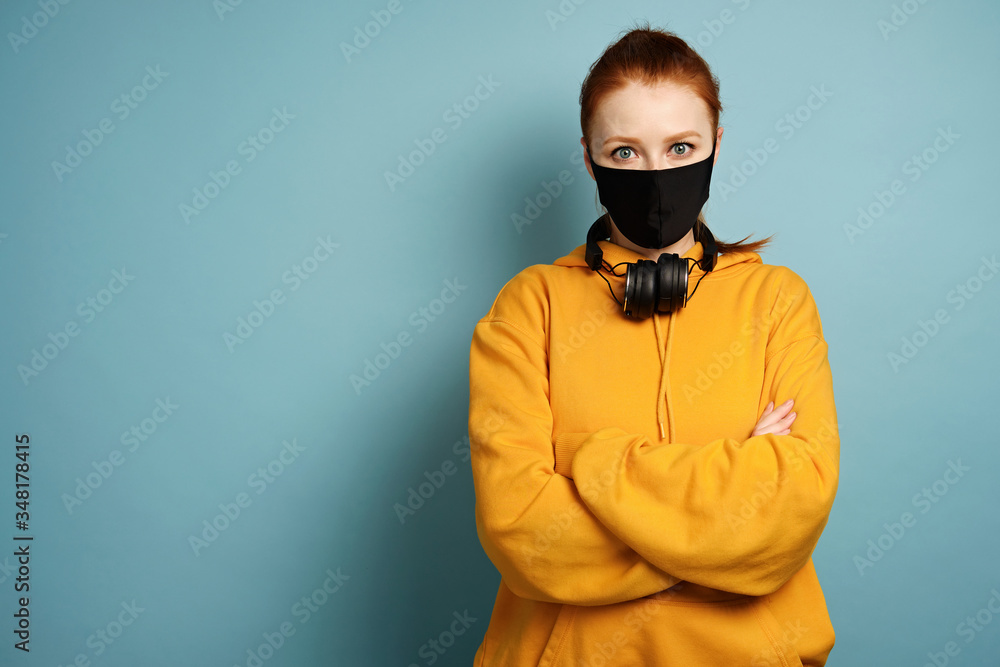 A girl in a yellow hoodie stands on a blue background in a black protective mask crossing arms on chest A girl in a yellow hoodie stands on a blue background in a black protective mask crossing arms on chest