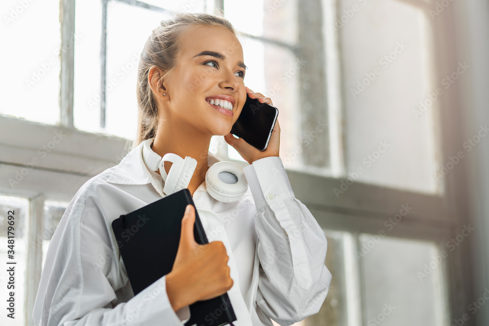 Smiling girl talking on smartphone, holding black notepad. Beautiful girl use mobile phone for work, call clients Smiling girl talking on smartphone, holding black notepad. Beautiful girl use mobile phone for work, call clients