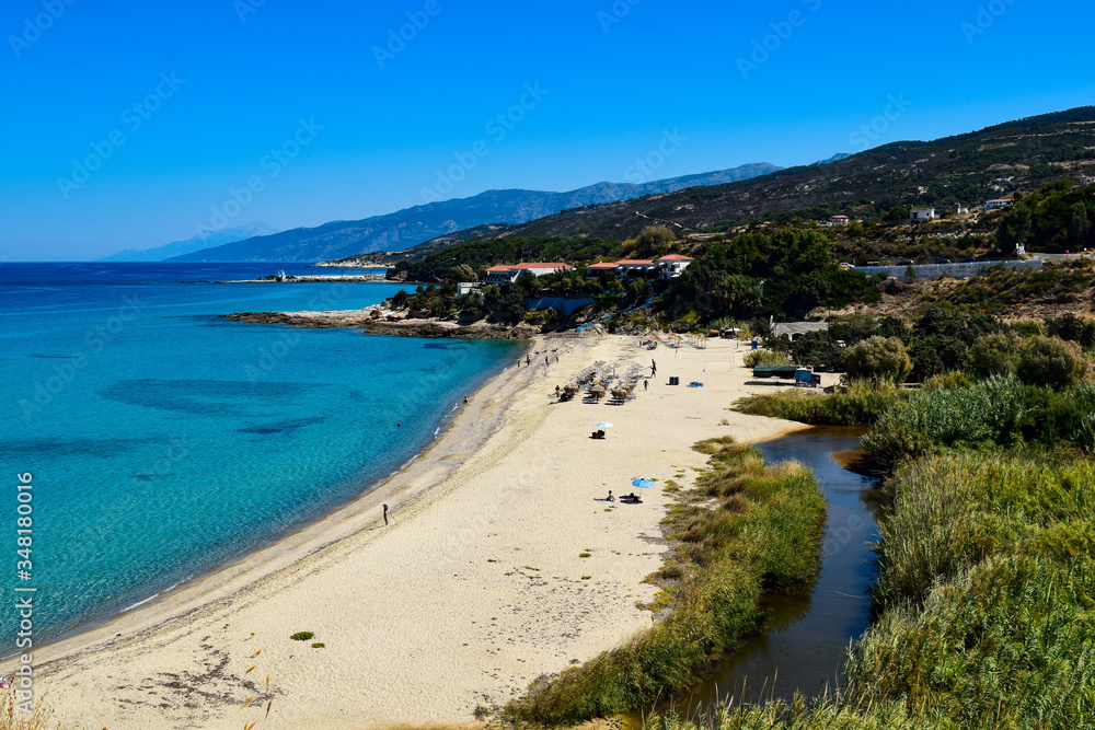 The Livadi Beach at the Ikaria island in a quiet summer day with blue clear water and nature behind, Ikaria, Greece. Little beach lagoon next to the shore.