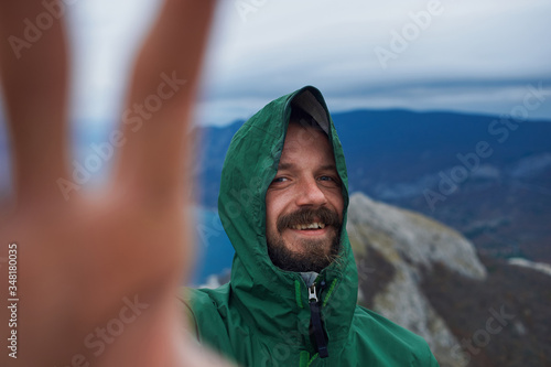 A young bearded traveler waves his hand at the camera on top of a mountain.