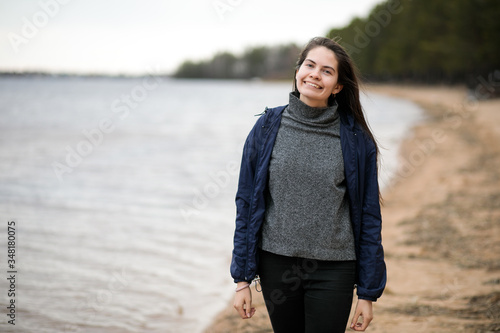 A beautiful smiling girl in a windbreaker stands on the sandy shore of a river or lake in early spring, look into the distance 