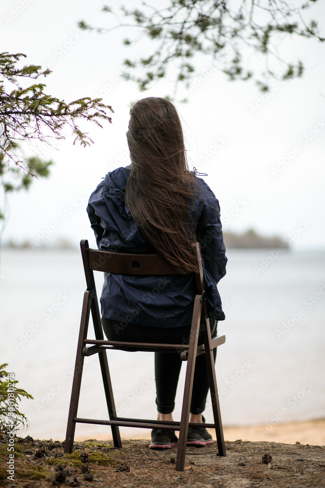 Fototapeta premium a young girl sits on a chair near a pond, against the background of a lake or river or sea, with her back to the camera, looks into the distance, admires the view and enjoys the fresh air 