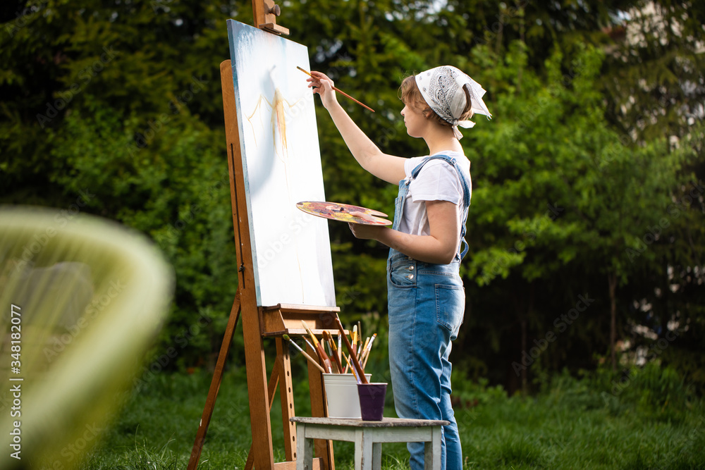 A young woman artist holds a brush and paints a picture on an easel in the rays of the sunset. The painter paints oil paintings in the garden