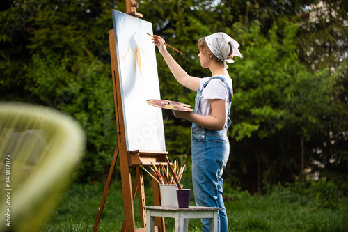Canvas Print A young woman artist holds a brush and paints a picture on an easel in the rays of the sunset