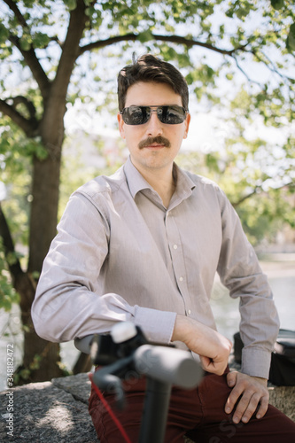 Portrait of man leaning on bike rudder. Serious man with moustache and sunglasses posing while sitting under tree. © Synergic Works OÜ
