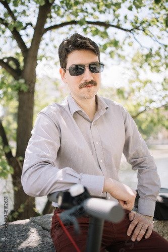 Handsome man with moustache and sunglasses posing under tree. Portrait of man with moustache wearing shirt and sunglasses while leaning on bike rudder.