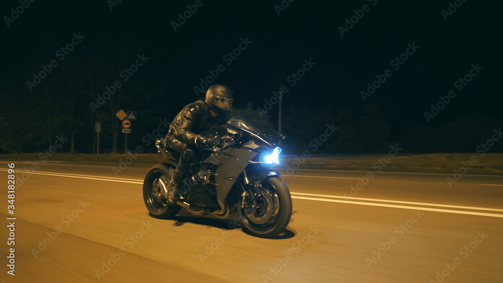 Young man in helmet riding fast on modern black sport motorbike at evening city street. Motorcyclist racing his motorcycle on night empty road. Guy driving bike. Concept of freedom and hobby. Close up