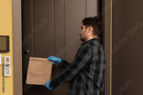Side view of handsome man in latex gloves holding package near open door