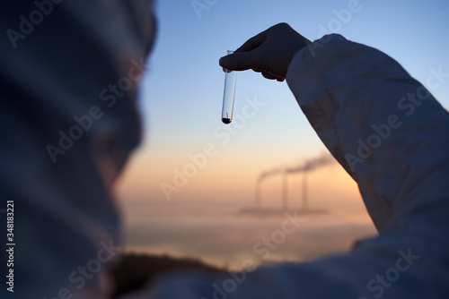 Cropped close-up snapshot of a scientist standing with his back to the camera, with his face to a view of smoking pipes of thermal power plant and looking at the glass flask with a sample. Ecology