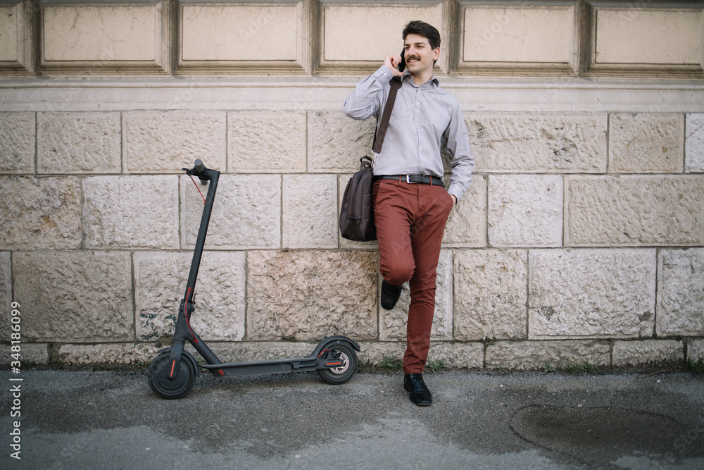Businessman posing while having conversation on the phone. Portrait of man with scooter leaning on wall while having a call and smiling. Businessman posing while having conversation on the phone. Portrait of man with scooter leaning on wall while having a call and smiling.