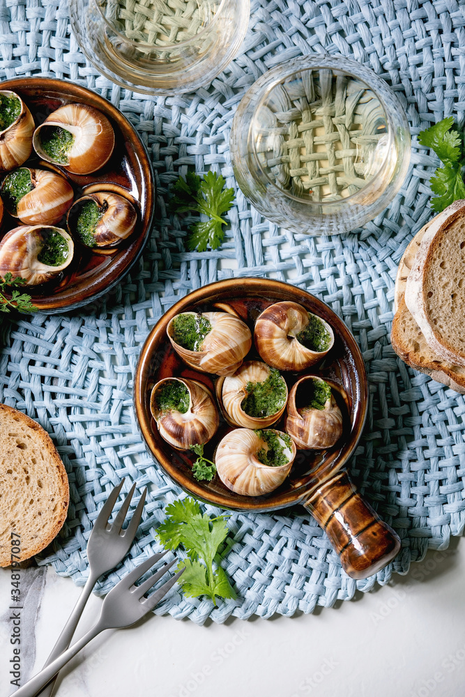 Escargots de Bourgogne - Snails with herbs butter, gourmet dish, in traditional ceramic pan with coriander, bread, glass of white wine on blue napkin over white marble background. Flat lay, space Escargots de Bourgogne - Snails with herbs butter, gourmet dish, in traditional ceramic pan with coriander, bread, glass of white wine on blue napkin over white marble background. Flat lay, space
