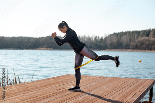 A fit young woman performs exercises with fitness elastic bands on a pier on the lake shore. The concept of a healthy lifestyle. Side view