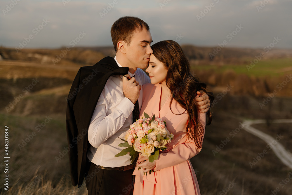 Naklejka premium Groom kisses bride on the forehead, against the background of a beautiful view of nature at sunset