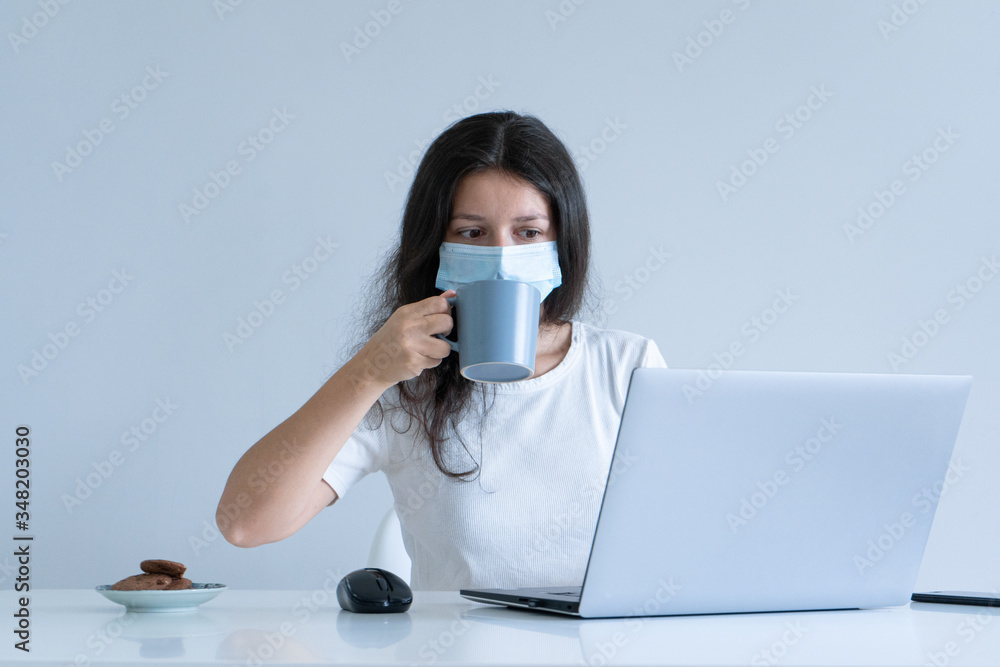 The girl works from home during quarantine. Corona virus pandemic. A girl in a surgical mask works on a laptop and drinks coffee. Distant work The girl works from home during quarantine. Corona virus pandemic. A girl in a surgical mask works on a laptop and drinks coffee. Distant work