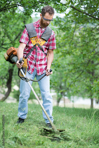 Determined young Caucasian man mowing grass in an orchard or back yard with a string trimmer