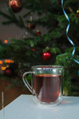 A glass cup of tea stands on a white table. In the background is a green Christmas tree decorated for New Year and Christmas.