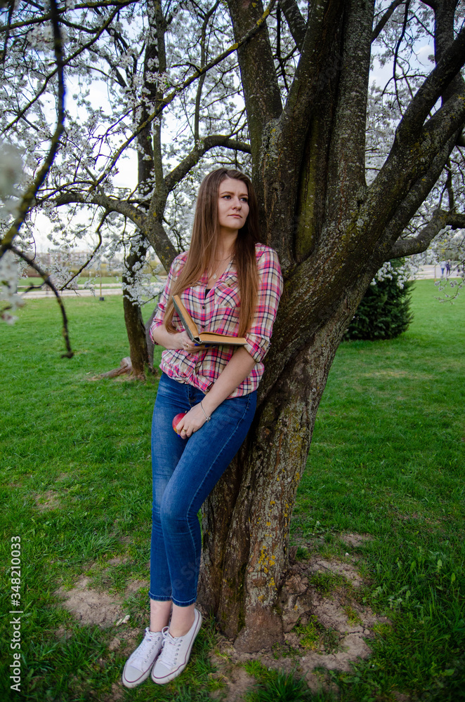 A girl stands near a tree and holds a book and an apple